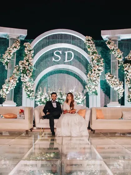 The couple seated on their magnificent reception stage. The scale of the design, with its towering arches and lush florals, creates a truly regal portrait of the newlyweds.