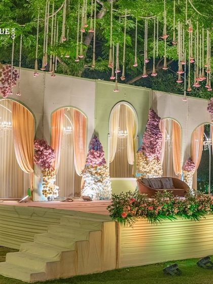 I designed this elegant reception stage with a backdrop of arched panels and soft pastel curtains. The hanging floral tassels and lush stage-front arrangements add layers of texture and romance.