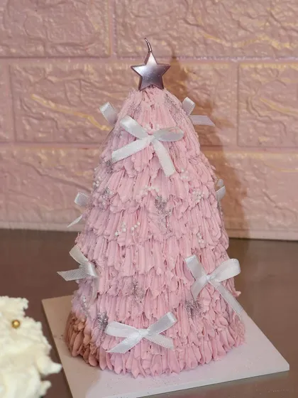 A cute and festive pink Christmas tree cake, covered in fluffy frosting, sparkles, and little white bows.