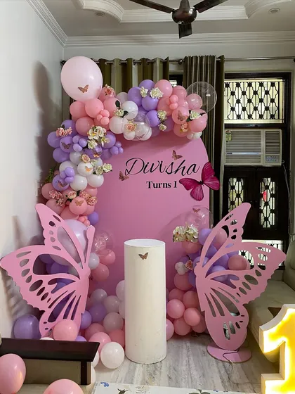 A lovely butterfly-themed first birthday decoration for a home setting. This design uses pink and purple balloons, large butterfly cutouts, and a custom pink backdrop.
