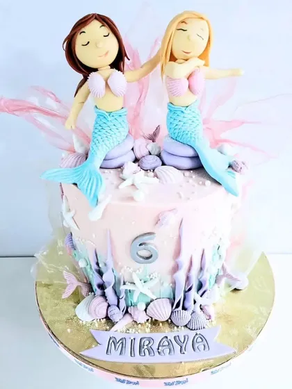 A charming mermaid cake for a sixth birthday. This design features two handcrafted mermaid friends sitting atop an ocean-themed cake, complete with edible shells and underwater foliage.