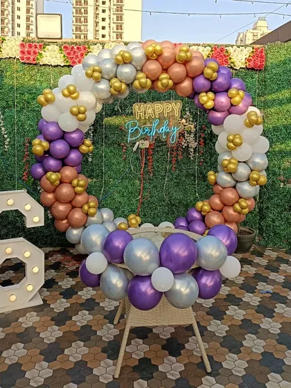 An outdoor birthday setup with a multi-color balloon ring against a green wall. The decoration includes a 'Happy Birthday' neon sign and a large light-up number '6'.
