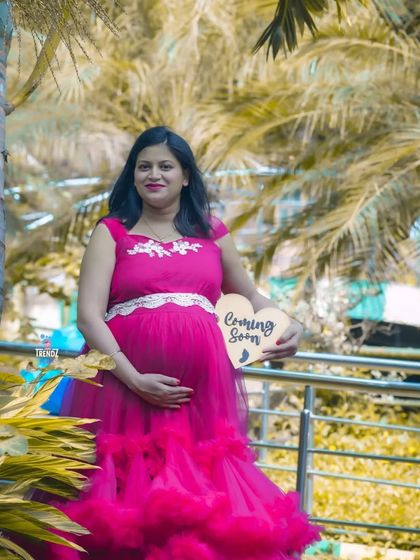 A lovely outdoor portrait of a mom-to-be holding a "Coming Soon" sign. The vibrant pink gown and the tropical foliage create a cheerful and warm announcement photo.