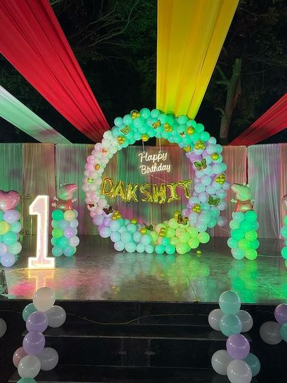 A large stage setup for an outdoor first birthday party. The decor includes a colorful fabric canopy, a balloon ring backdrop, and light-up balloon columns.