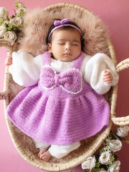 A peaceful portrait of a baby sleeping in a basket, dressed in a lovely purple outfit and surrounded by white roses.