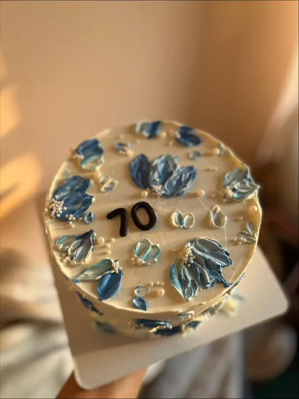 A close-up of a 70th birthday cake decorated with blue and white palette knife flowers. The chocolate sponge is filled with salted caramel and covered in vanilla buttercream.