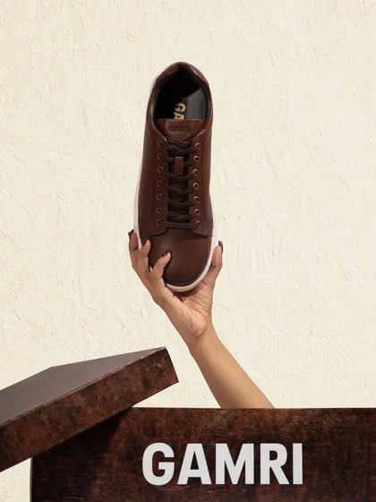 A creative shot of a hand lifting a brown leather sneaker out of its box. This "unboxing" style image creates anticipation and highlights the product in a dynamic way.