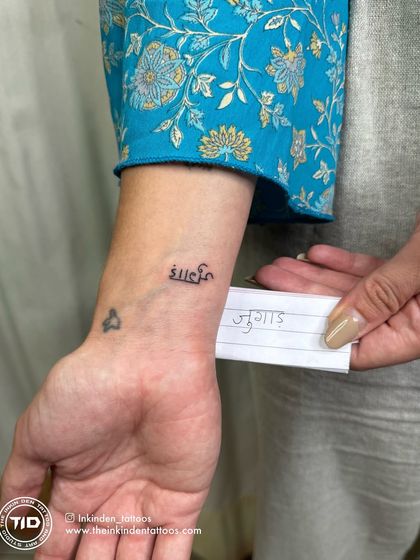 A close-up of the 'Shanti' script tattoo on the client's wrist, with the handwritten note for 'Jugaad' in the background.