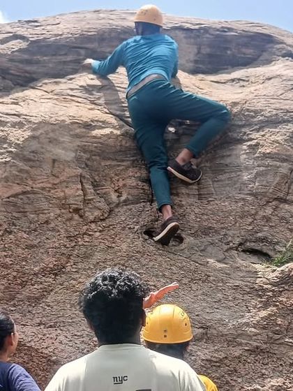 A participant in full gear practices his ascent on a large boulder in Ramanagara, with fellow campers watching and learning.