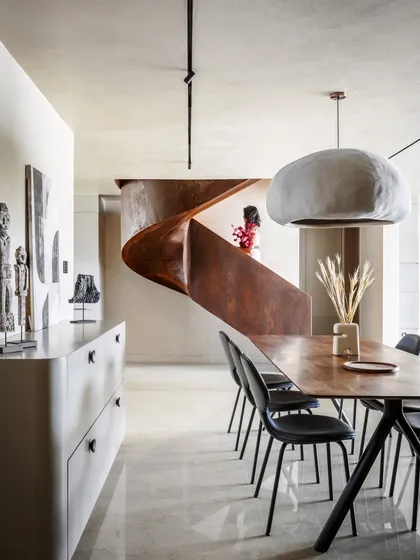 The dining area is positioned to offer a view of the magnificent Corten steel staircase. The simple wooden table and modern chairs are complemented by a large, bespoke pendant light, creating an elegant setting for meals.