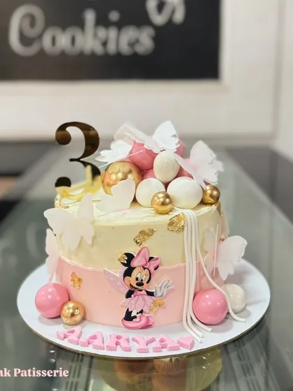 An elegant Minnie Mouse cake featuring a delicate character cutout, butterflies, and decorative spheres. The pink and gold color scheme adds a touch of magic.