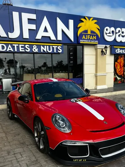 A vibrant red Porsche 911 GT3 with a racing stripe parked in front of the Ajfan Dates & Nuts store, adding a splash of high-speed color to our storefronts.