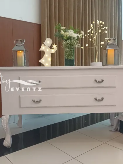 We provide decorative furniture and props for your event, like this white console table for a baptism ceremony. It's decorated with lanterns, a light-up angel, and a vase of white lilies.