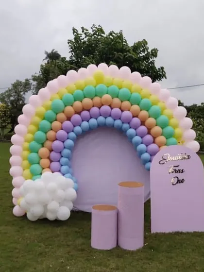 A beautiful pastel rainbow balloon arch set up in a large garden. This simple yet impactful decoration is perfect for a daytime outdoor party for a child's first birthday.