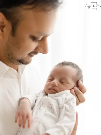 A father holds his newborn close, looking down with a gentle smile. The soft, natural light enhances the tenderness of the moment.
