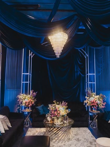 A chic lounge area for guests at the Sangeet, featuring deep blue velvet sofas, modern white structures, and elegant chandeliers.