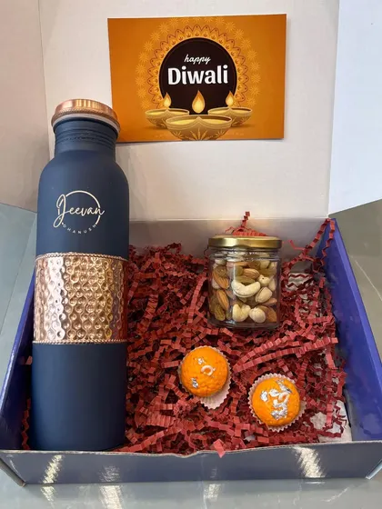 A simple and elegant Diwali gift box. It contains a personalized hammered copper bottle, a jar of dry fruits, and traditional motichoor ladoos, complete with a "Happy Diwali" card.