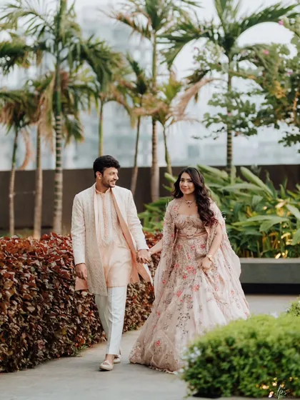Aayushi and Ameya walk hand-in-hand through a lush garden path. The soft, natural light and their coordinated pastel outfits create a romantic and serene portrait.