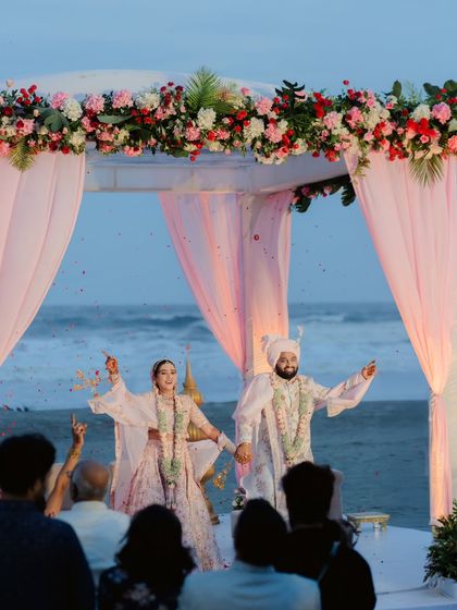 The couple celebrates at their beachside mandap, surrounded by guests. The soft pink drapes and floral arrangements create a romantic setting against the evening sky.