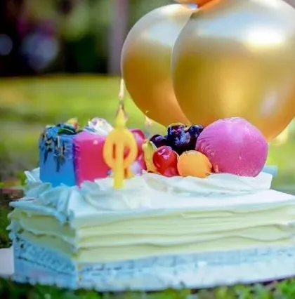 A colorful birthday cake topped with fruit, a candle, and golden balloons, ready for an outdoor celebration.