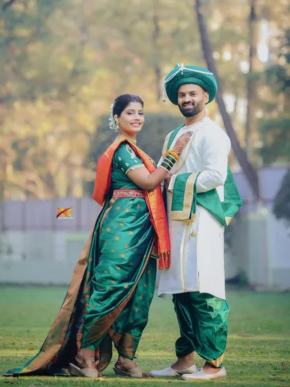 A classic full-length portrait of the couple in their traditional Maharashtrian wedding attire. The bride's green Nauvari saree and the groom's coordinated outfit look perfect in this outdoor setting.