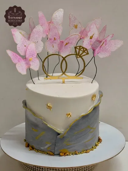 A beautiful two-tier engagement cake in a grey and white marble design with gold leaf accents. The top is decorated with delicate pink wafer paper butterflies and a golden ring topper, making it both modern and romantic.