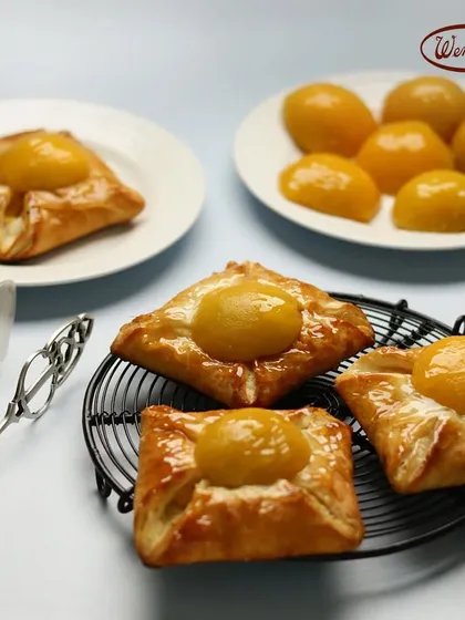 Our Peach Danish pastries, fresh and golden. These feature a flaky puff pastry base with a creamy cheese filling and a sweet peach half on top.
