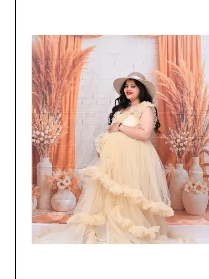 A stylish solo portrait featuring a beautiful cream-colored tulle gown and a wide-brimmed hat. The warm, peachy tones of the pampas grass backdrop create a cohesive and fashionable look for this Boho-chic maternity photo.