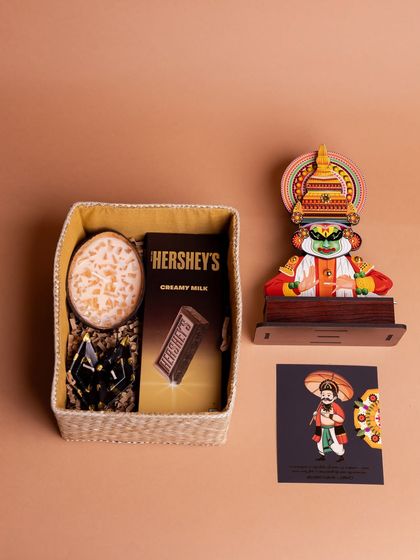 The 'Onakazhcha' hamper deconstructed to show its contents. This gift set includes a Kathakali standee, Hershey's chocolate, a scented candle, and a Mahabali card, perfect for a modern Onam gift.