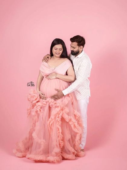 A full-length view of this gorgeous couple's portrait. The off-shoulder pink gown with its cascading ruffles is pure romance, and his simple white shirt keeps the focus on her and the baby bump.