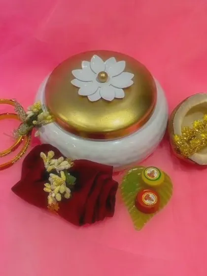 This elegant white and gold metal box features a lotus handle and holds a complete tamboola set. Inside, your guests will find a folded blouse piece, bangles, a half coconut, and a leaf-shaped haldi-kumkuma container, making it a cherished keepsake.