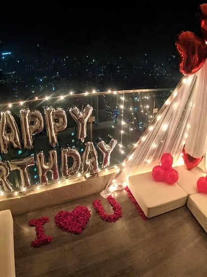 A romantic birthday surprise on a balcony, featuring a small teepee, 'Happy Birthday' foil balloons, and a personal message. The 'I ❤️ U' made from rose petals adds a heartfelt touch.