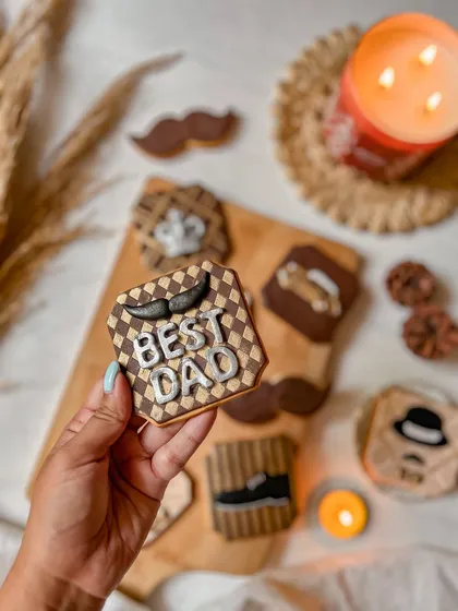 A hand holding up the "Best Dad" cookie from the Father's Day set, showing the detailed checkerboard pattern and icing work. It's a personal and stylish way to celebrate him.