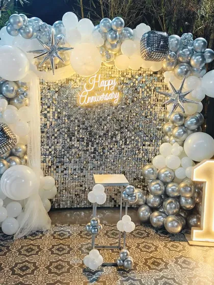 A stunning first-anniversary celebration featuring a silver sequin shimmer wall. The setup is framed by a chrome and white balloon arch, with a "Happy Anniversary" neon sign and a light-up number '1' for a touch of sparkle.