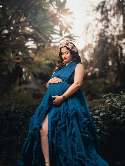 A close-up portrait of a mother-to-be in a lush green setting, wearing a deep blue gown and a floral crown. Her serene expression captures a moment of quiet reflection.