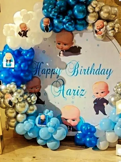This Boss Baby first birthday setup for Aariz featured a circular backdrop with multiple characters, a light-up number '1', and baby blocks spelling out his name, all framed by a blue and silver balloon arch.