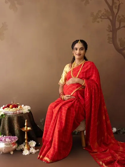 A classic seated portrait of a mom-to-be in a red saree, embodying grace and tradition in a studio setting.