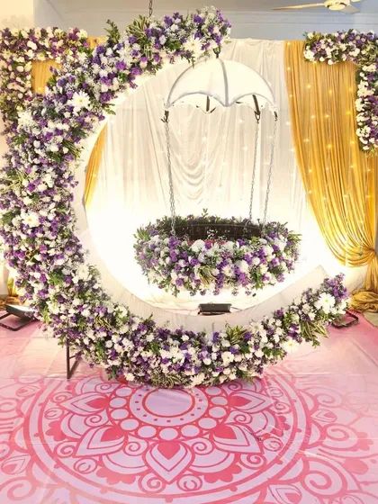 I create elegant stages for naming ceremonies, like this one with a crescent moon floral frame. The cradle is also decorated with matching purple and white flowers, and the backdrop uses sheer and gold curtains for a traditional and beautiful seemantham event.