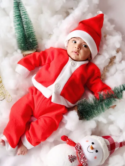 An aerial shot of a baby in a Santa outfit lying in a snowy landscape with a snowman and tiny trees.