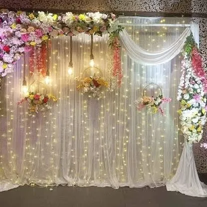 A beautiful and cost-effective backdrop using a curtain of fairy lights and simple white drapes. We added a floral runner at the top, hanging floral baskets, and exposed bulbs to create a warm and inviting photo corner.