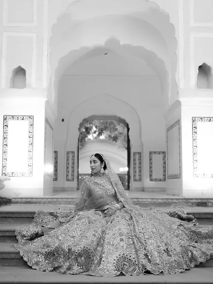 A dramatic black-and-white version of the regal bridal portrait, emphasizing the form and texture of her elaborate lehenga against the architectural backdrop.
