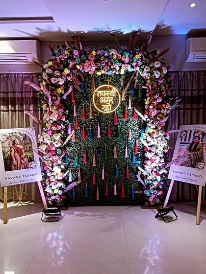 A festive backdrop for a Navanu Tapasvi event. The design includes a dense floral arch over a green wall with colorful tassels and personalized photo stands on either side.
