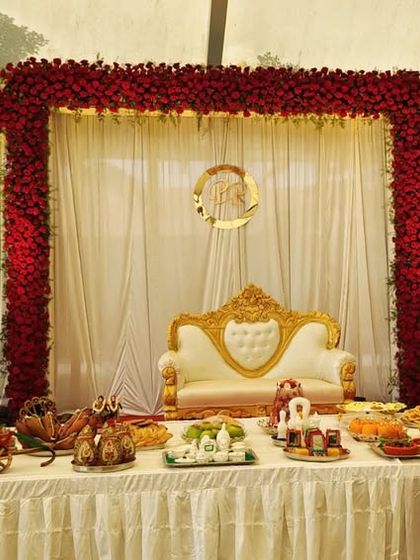 A traditional engagement stage setup featuring a backdrop of red roses and white drapes, with a table laden with customary sweets and offerings.