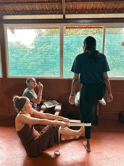 We teach you to see the body and its needs. A student learns to use straps to support another in a hamstring stretch, focusing on safe alignment and communication to facilitate a deeper release.