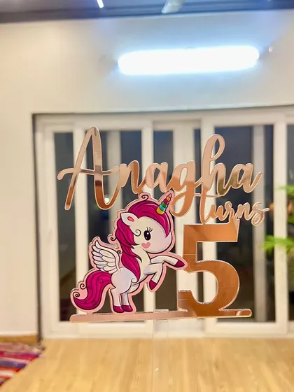 The "Anagha turns 5" unicorn topper held up to show its full design and the clear acrylic stick that holds it securely on the cake.