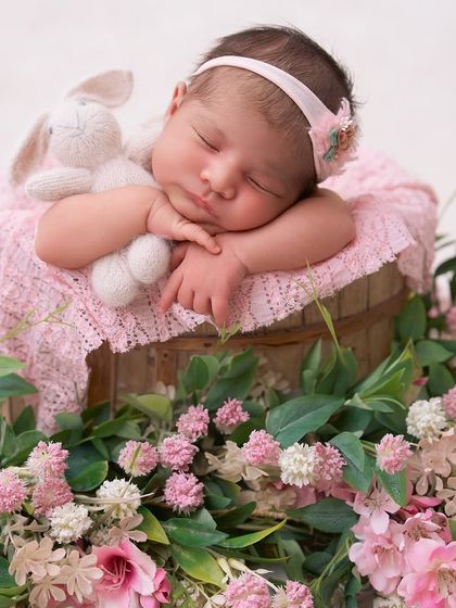 A sweet baby girl cuddles a knitted bunny while sleeping in a wooden bucket surrounded by a ring of fresh pink and white flowers and greenery.