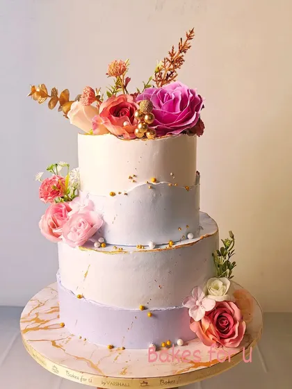 An elegant three-tier engagement cake with a soft lavender and white color palette. The cake is adorned with fresh flowers and gold leaf, creating a romantic and sophisticated look.