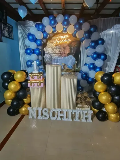 We travel for outside decorations, like this first birthday event in Hoovinamadu. The setup includes a blue and white balloon arch, gold and black balloon columns, and the name 'NISCHITH' in letters.