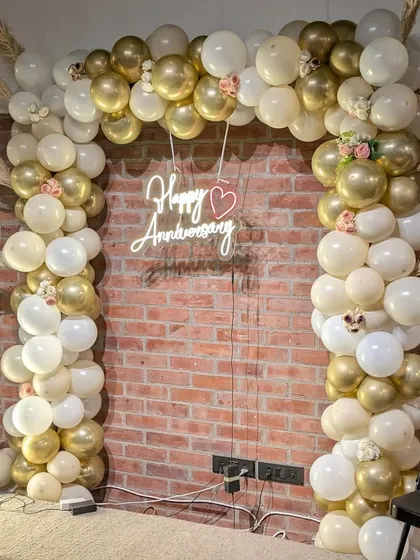 I design elegant backdrops for milestone celebrations like this 25th anniversary. A custom balloon arch in a sophisticated gold and white palette, paired with a 'Happy Anniversary' neon sign, creates a beautiful photo corner.