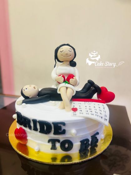 A funny and cute 'Bride to Be' cake. It features a fondant bride relaxing while the groom lies down, with a calendar marking the big day.